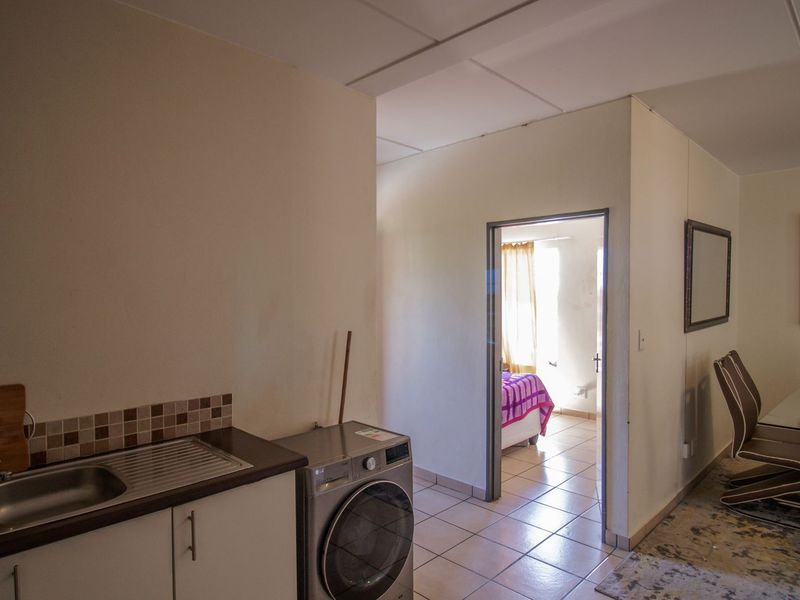 Open plan living area featuring a compact kitchen corner with sink and tiled backsplash, leading into an adjacent bedroom space with tiled flooring throughout.