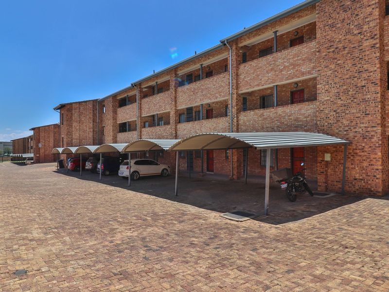 Exterior view of a multi-story brick apartment building featuring covered parking bays and a paved driveway area.