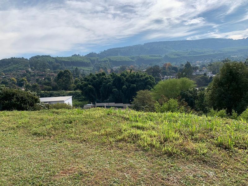 A scenic view overlooking a sloped residential plot with lush greenery and distant mountain landscape.
