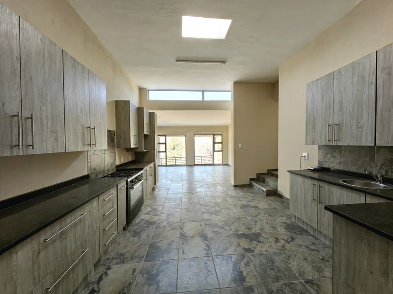 Spacious open-plan kitchen featuring modern wood-look cabinetry, dark stone countertops, and tiled flooring, leading into a bright living area with large windows.