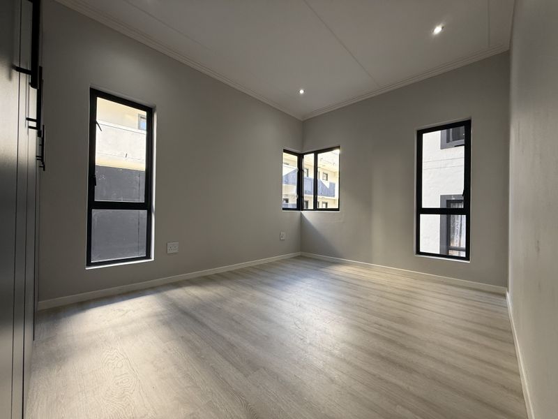 A bright, modern, and empty bedroom featuring light-toned laminate flooring, recessed downlights, and built-in cupboards with sleek black hardware.
