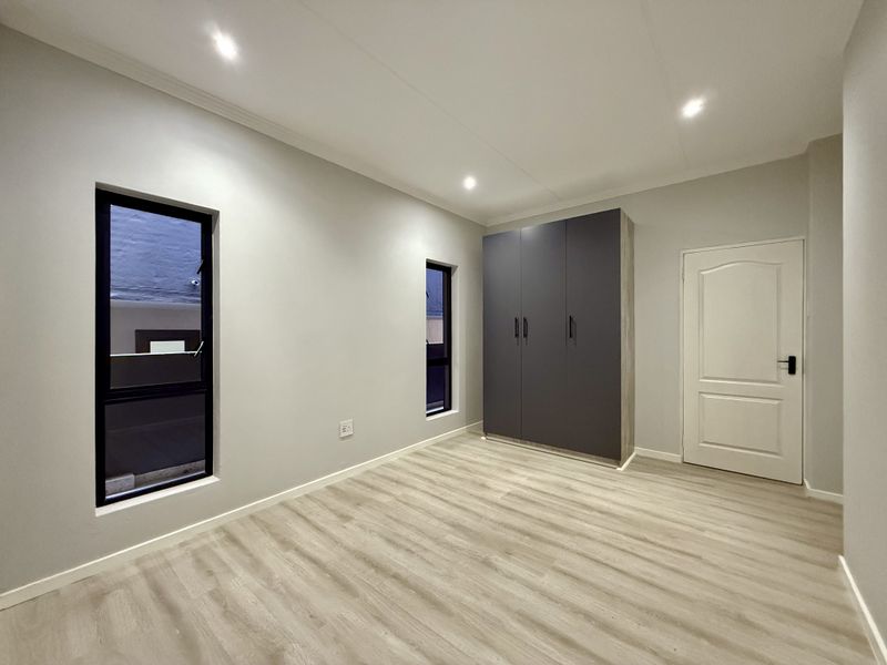 A bright, modern bedroom featuring light laminate flooring, recessed downlights, and a sleek dark grey built-in wardrobe.