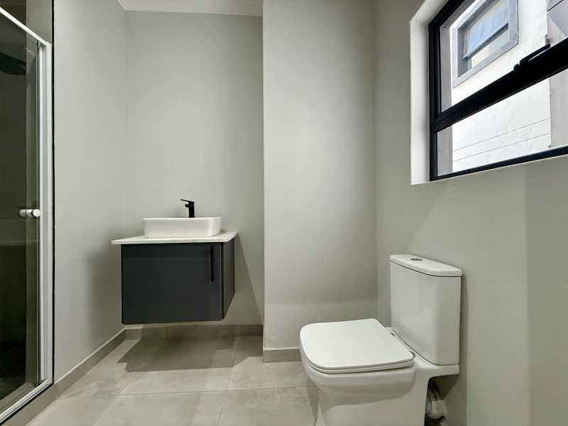A modern, minimalist bathroom featuring a floating vanity with a vessel sink, black fixtures, a toilet, and a glass-enclosed shower.