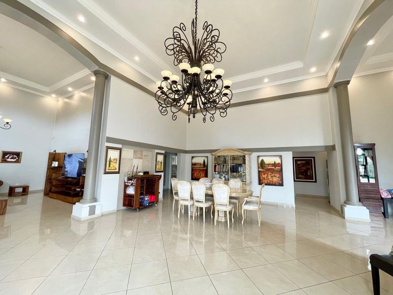 Spacious open-plan living and dining area featuring high ceilings, decorative columns, polished tile flooring, and an elegant central chandelier.