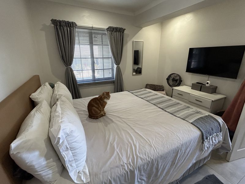 A well-maintained bedroom featuring tiled flooring, a window with venetian blinds and curtains, and a wall-mounted television.