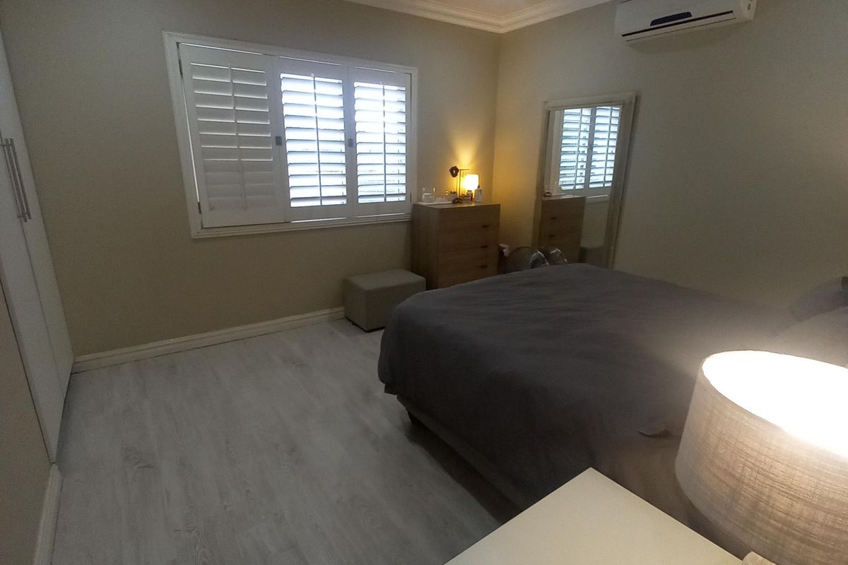 A well-maintained bedroom featuring light-toned laminate flooring, plantation shutters, built-in cabinetry, and a wall-mounted split-system air conditioner.