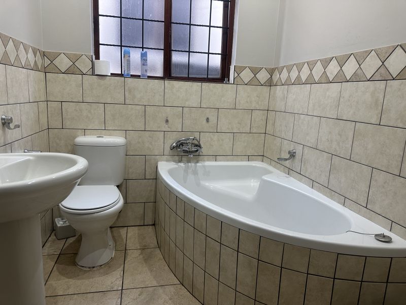A functional bathroom featuring a corner bathtub, pedestal basin, and toilet, finished with neutral wall and floor tiling.