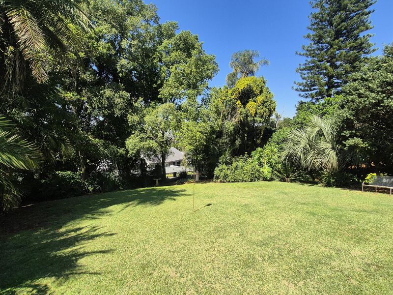 A spacious, well-maintained garden featuring a large manicured lawn surrounded by mature trees and established greenery.