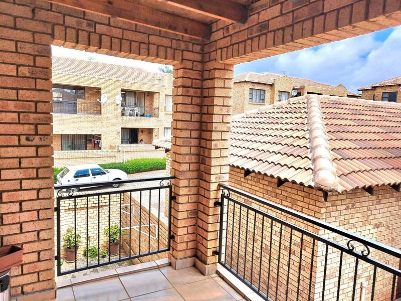 A covered balcony featuring face-brick pillars, tiled flooring, and black metal railings, overlooking the apartment complex grounds.