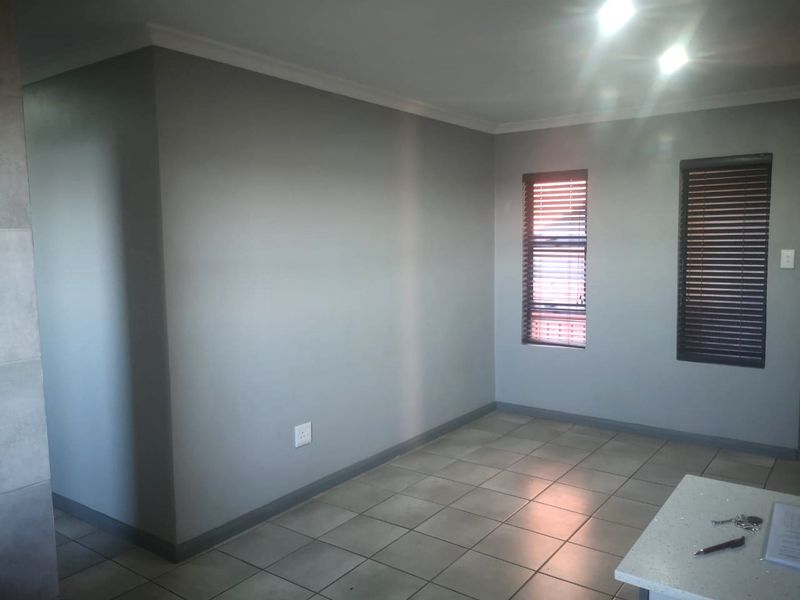 A bright, empty living area featuring neutral grey walls, tiled flooring, and fitted window blinds, offering a clean and versatile space.