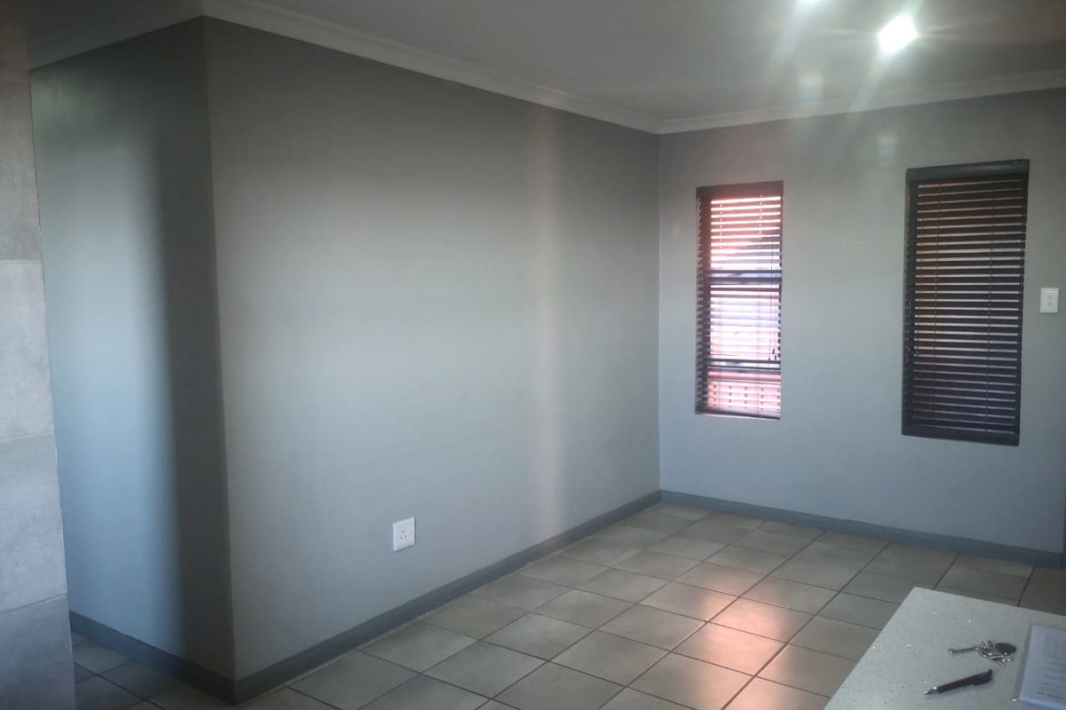 A bright, empty living area featuring neutral grey walls, tiled flooring, and fitted window blinds, offering a clean and versatile space.
