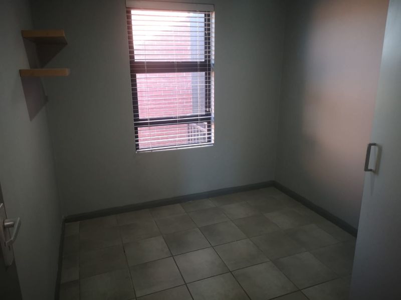 A compact, empty bedroom featuring tiled flooring, a window with fitted blinds, and wall-mounted shelving.
