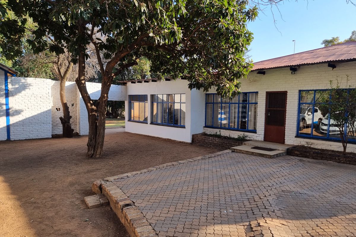 Rear exterior view of a small holding property featuring painted brick walls, steel-framed windows, and a paved courtyard area under mature trees.