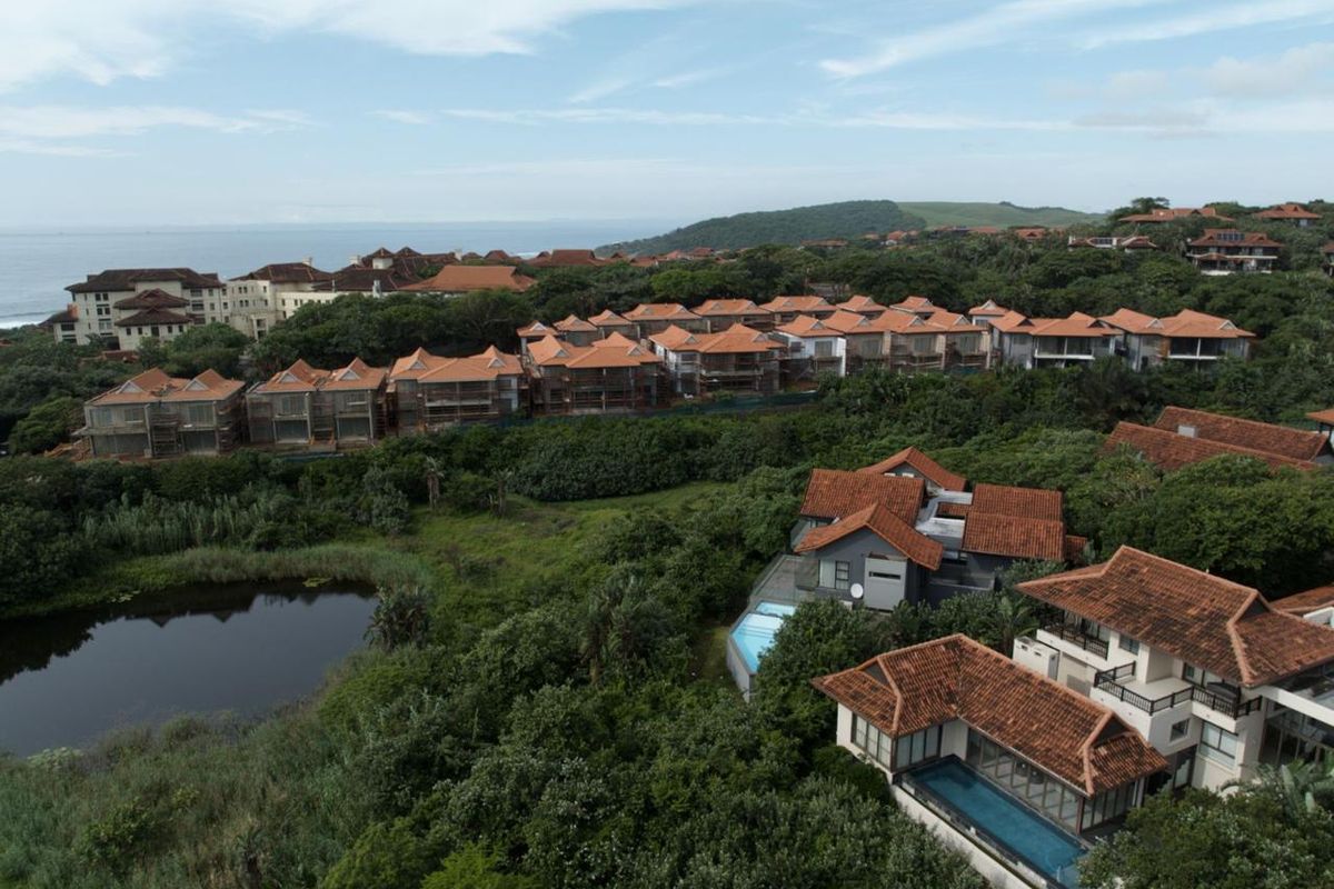 Aerial view of a coastal residential estate featuring multiple villas with tiled roofs, private swimming pools, and lush surrounding greenery.