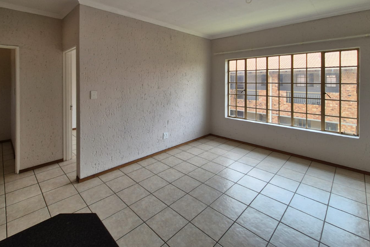 An empty, well-lit living room featuring neutral tiled flooring, textured walls, and a large window with security bars.