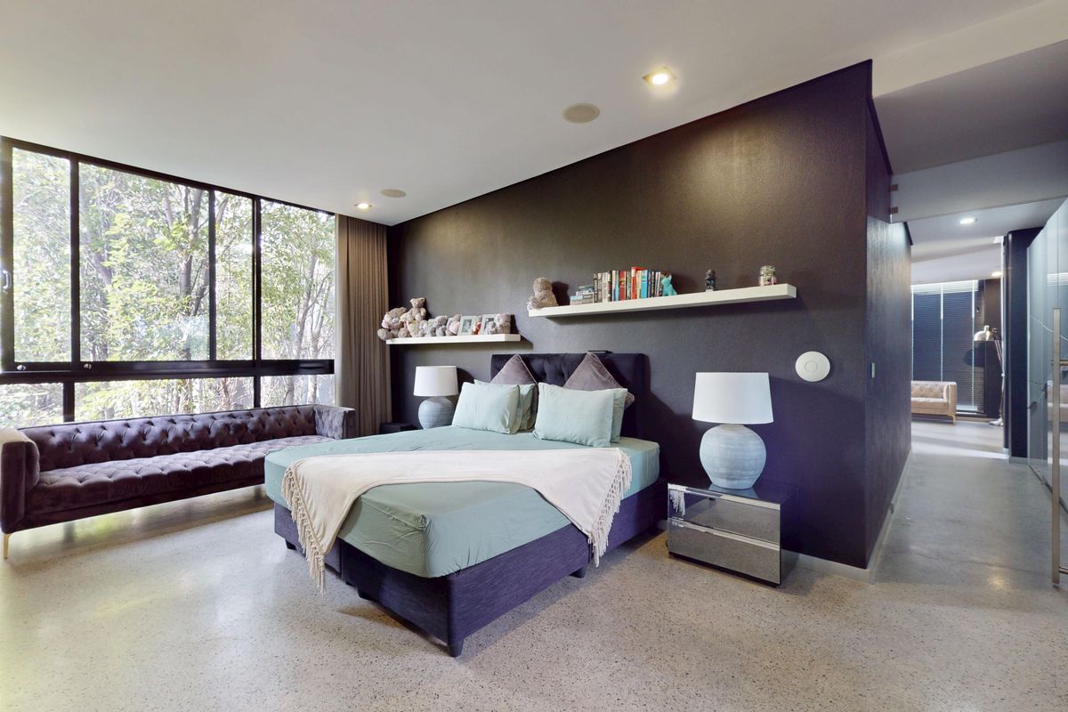 Spacious modern bedroom featuring polished concrete floors, large floor-to-ceiling windows with natural views, and dark accent wall with integrated floating shelving.