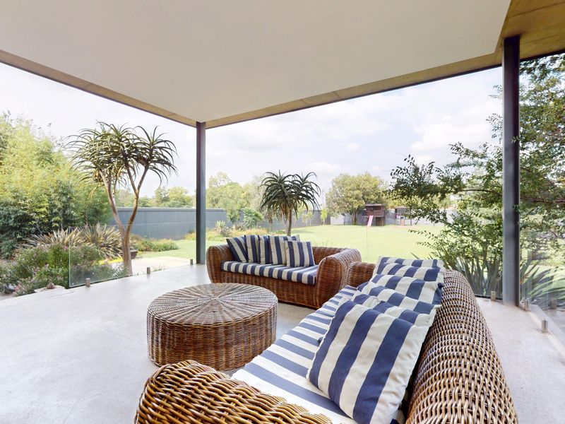 A spacious covered patio featuring concrete flooring and a modern glass balustrade, overlooking a well-maintained garden area.