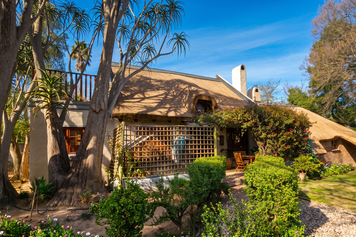 Rear view of a charming thatched-roof house featuring a private garden, mature trees, and a small patio area.