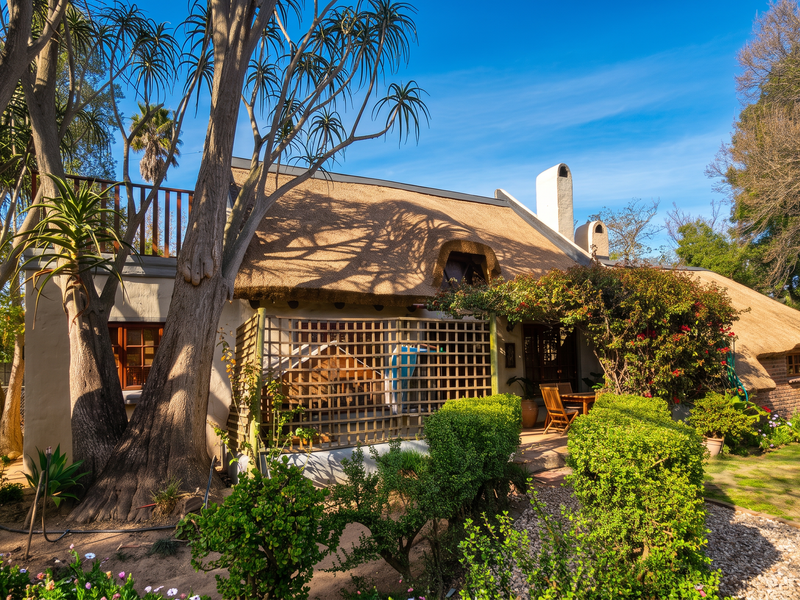 Rear view of a charming thatched-roof house featuring a private garden, mature trees, and a small patio area.