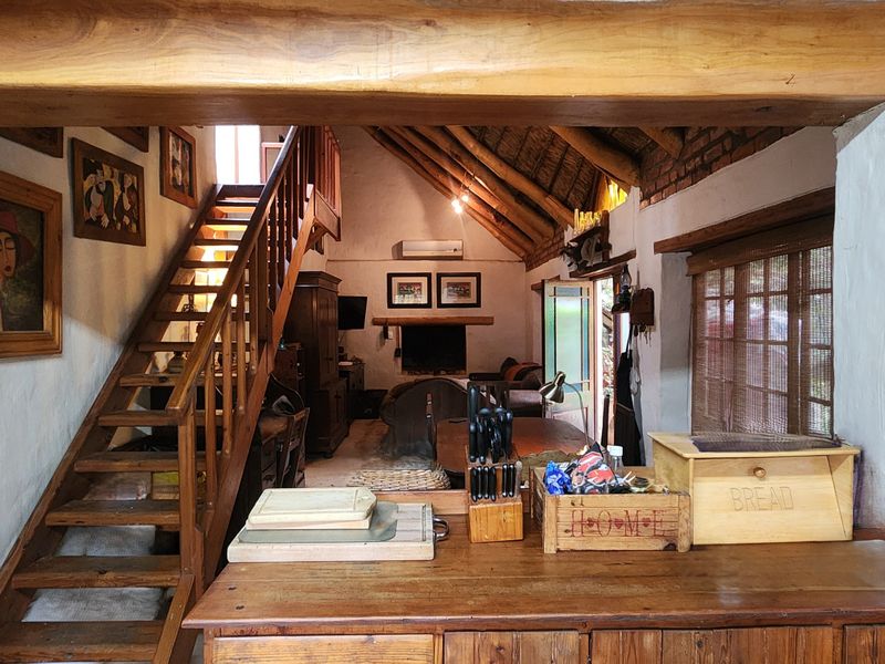 Open-plan living area featuring exposed timber beams, a thatched roof, a central fireplace, and a timber staircase leading to an upper level.