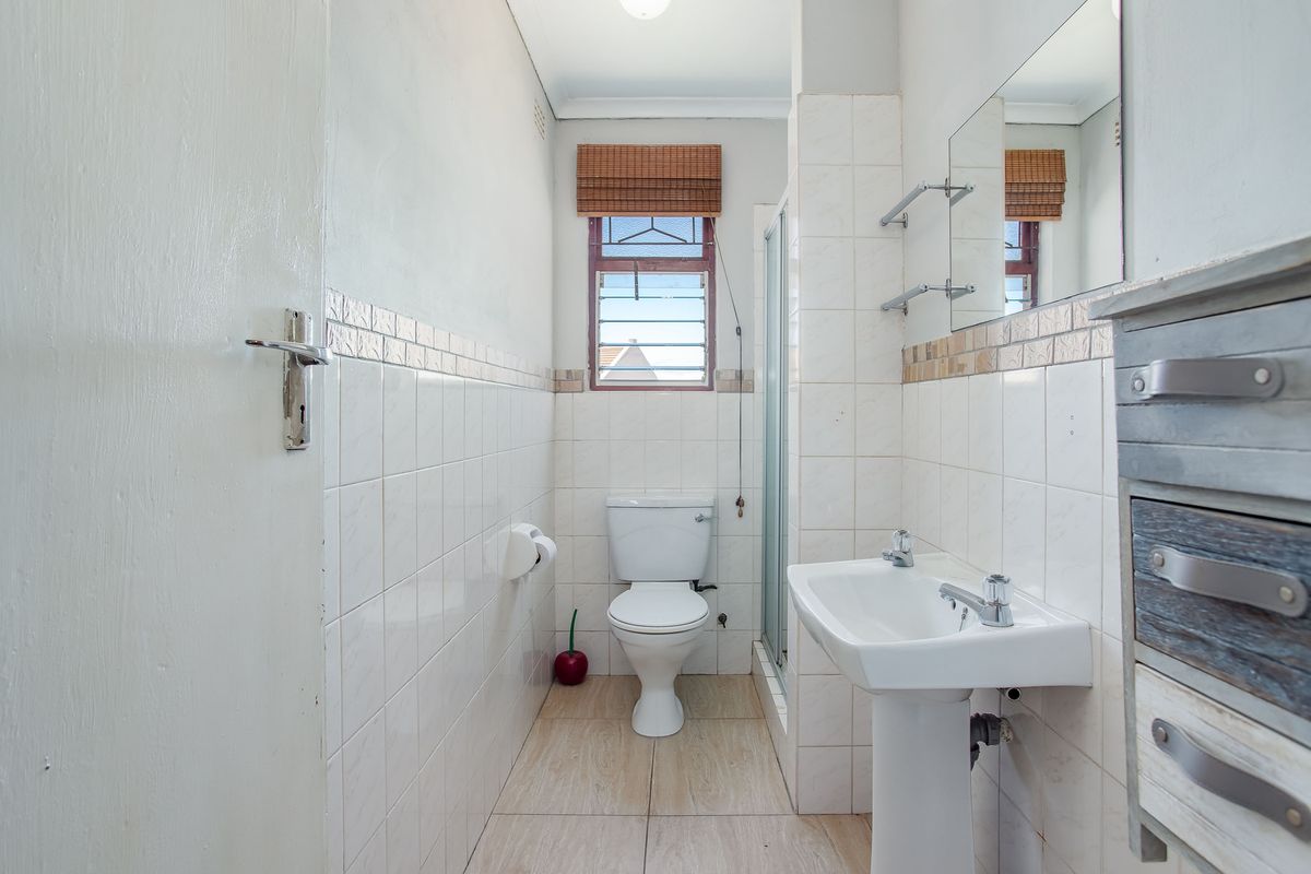 A functional bathroom featuring a pedestal washbasin, toilet, and shower enclosure with tiled finishes and natural light from a louvered window.