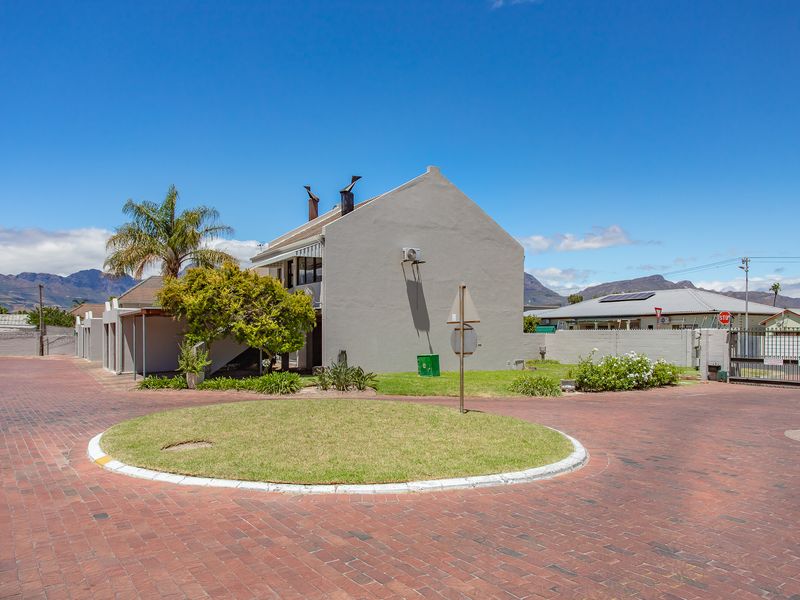 Exterior view of the apartment complex featuring a brick-paved driveway, landscaped roundabout, and secure perimeter fencing with mountain views in the background.