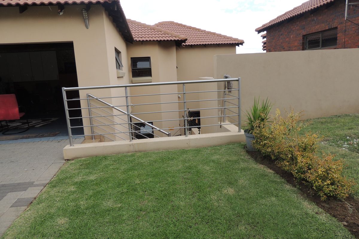 Rear exterior view featuring a paved patio area, stainless steel balustrade, and a well-maintained lawn with boundary walling.