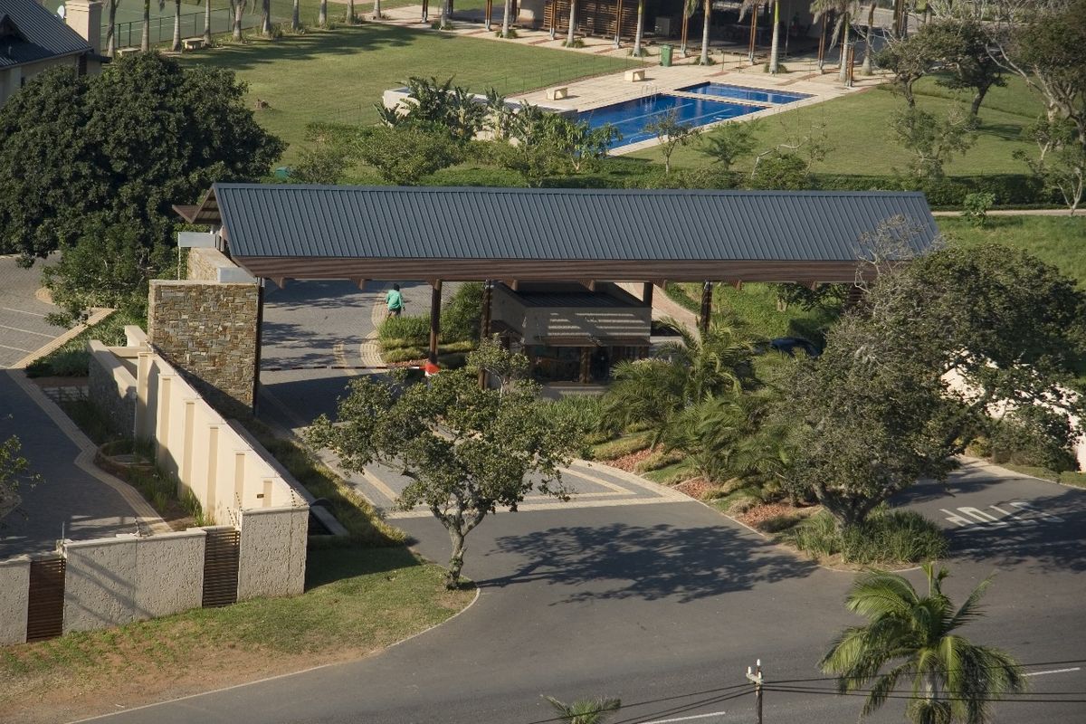 Aerial view of a secure residential estate entrance featuring a modern gatehouse, paved driveway, and landscaped surroundings with a nearby swimming pool.