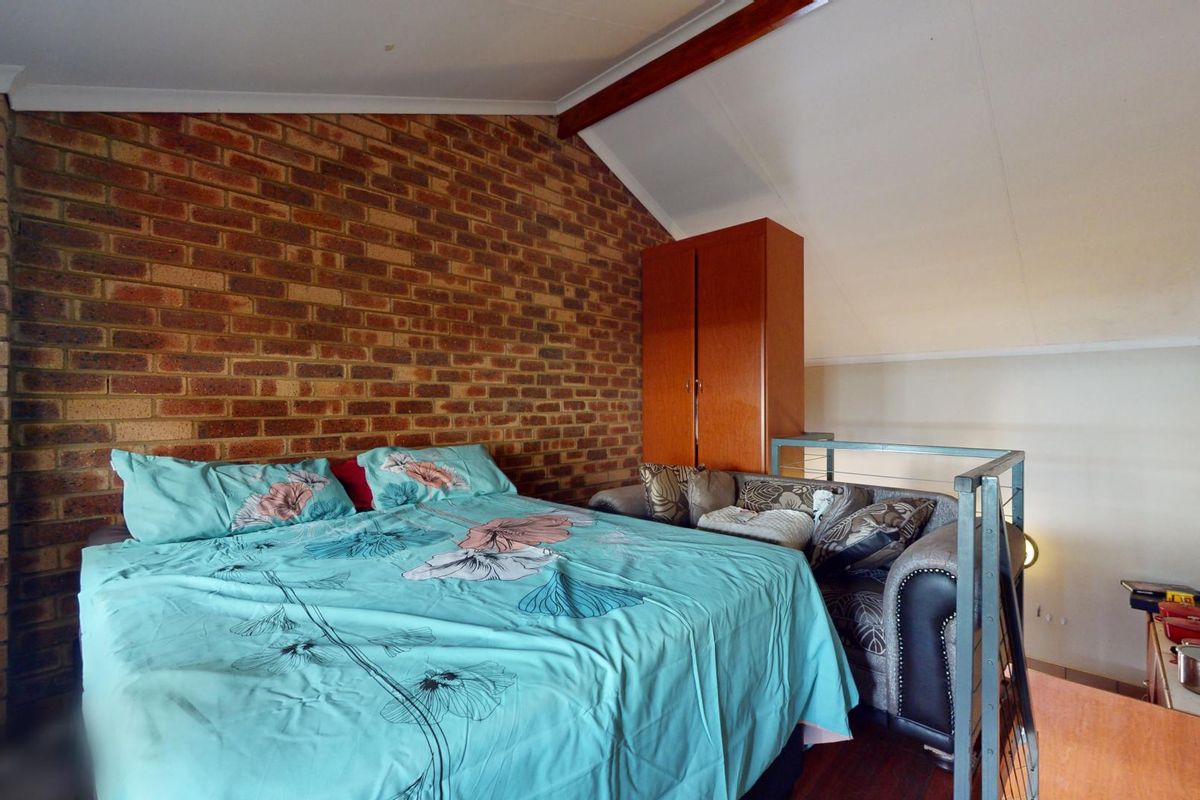 A loft-style bedroom featuring an exposed brick feature wall, vaulted ceiling with timber beam, and a metal safety balustrade overlooking the lower level.