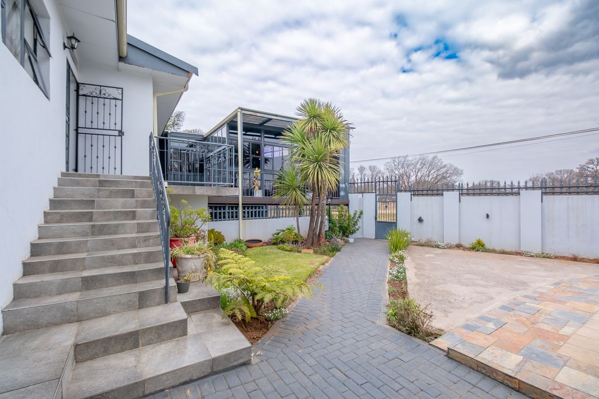 A well-maintained rear exterior area featuring a paved walkway, concrete staircase, landscaped garden, and a modern enclosed patio structure with metal railings.