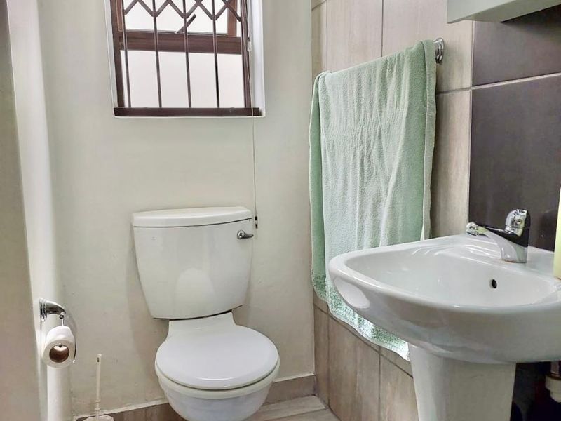 A compact bathroom featuring a pedestal basin, toilet, and neutral wall and floor tiling with a security-fitted window.