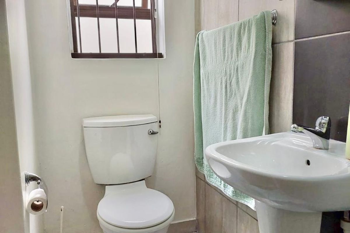 A compact bathroom featuring a pedestal basin, toilet, and neutral wall and floor tiling with a security-fitted window.