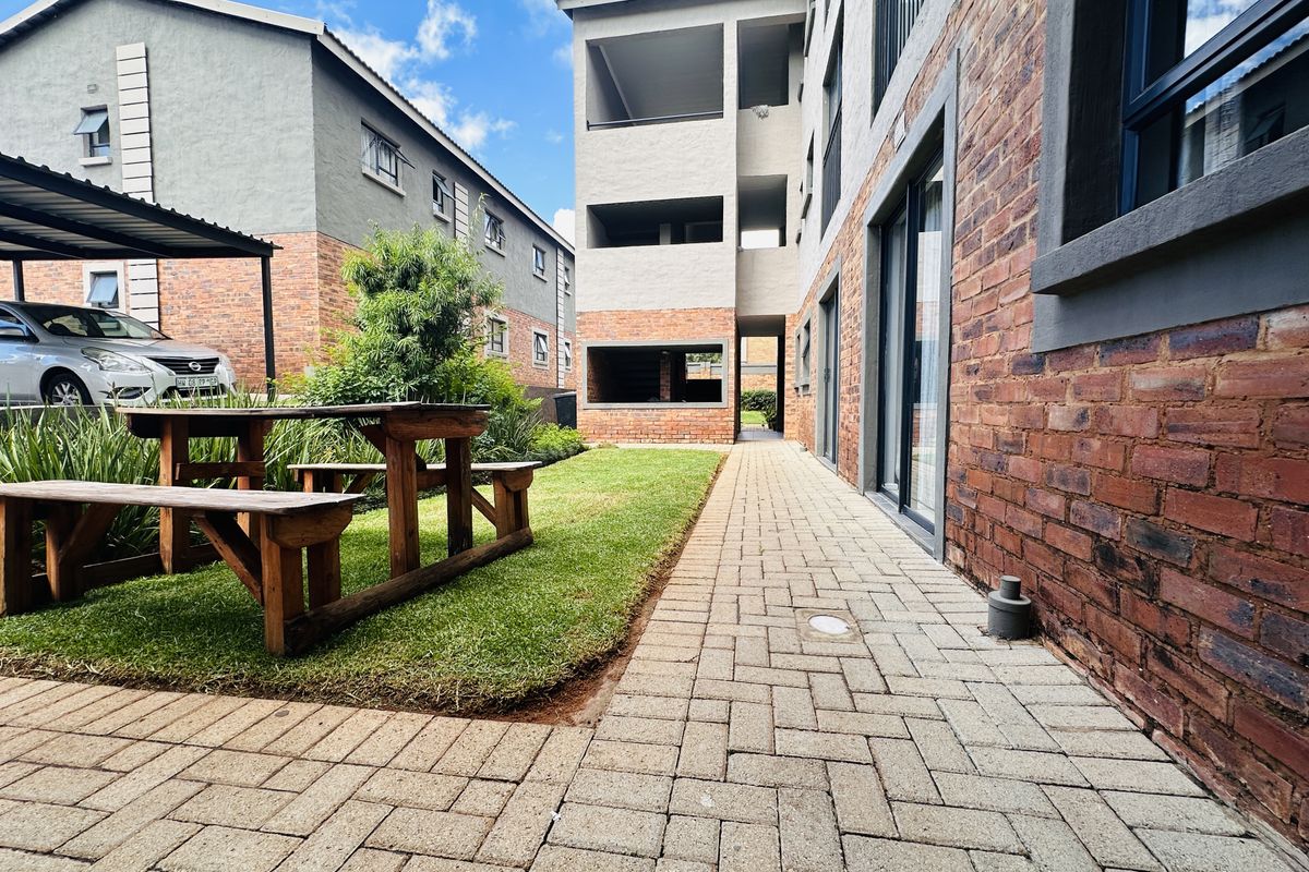 A well-maintained exterior rear courtyard area featuring a paved walkway, manicured lawn, and brick apartment building facade with sliding glass door access.