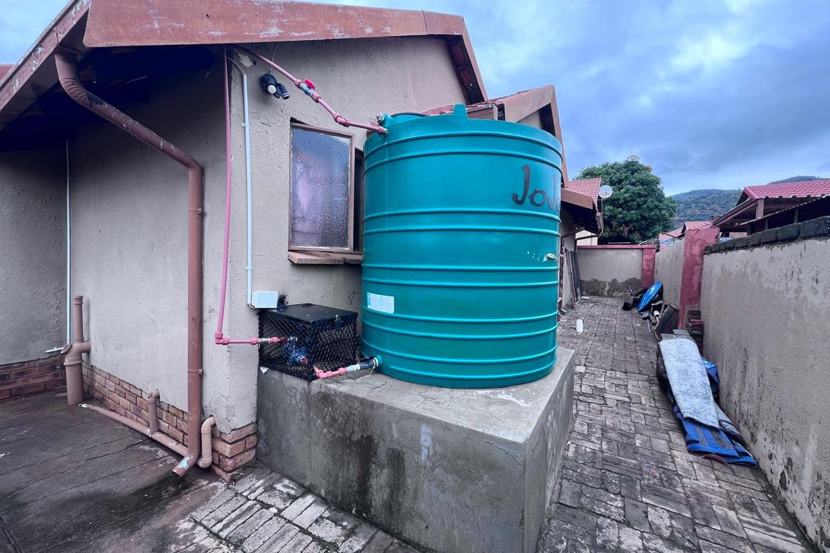 Rear exterior view showing a water storage tank on a concrete plinth, paved walkway, and boundary wall.