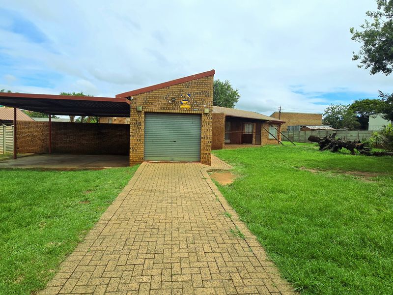 Front view of a brick house featuring a single garage, attached carport, paved driveway, and a large front garden area.