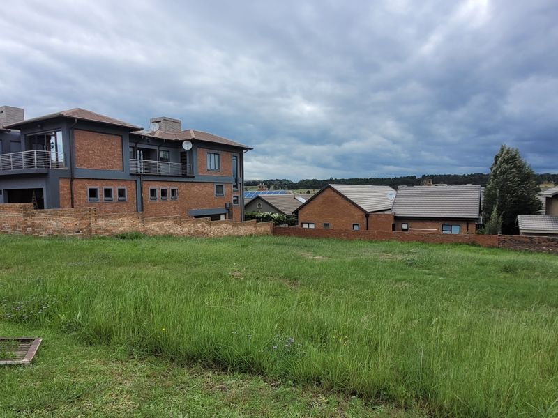A vacant residential plot featuring a grass lawn and brick boundary wall, situated in a developed neighborhood with surrounding residential properties.