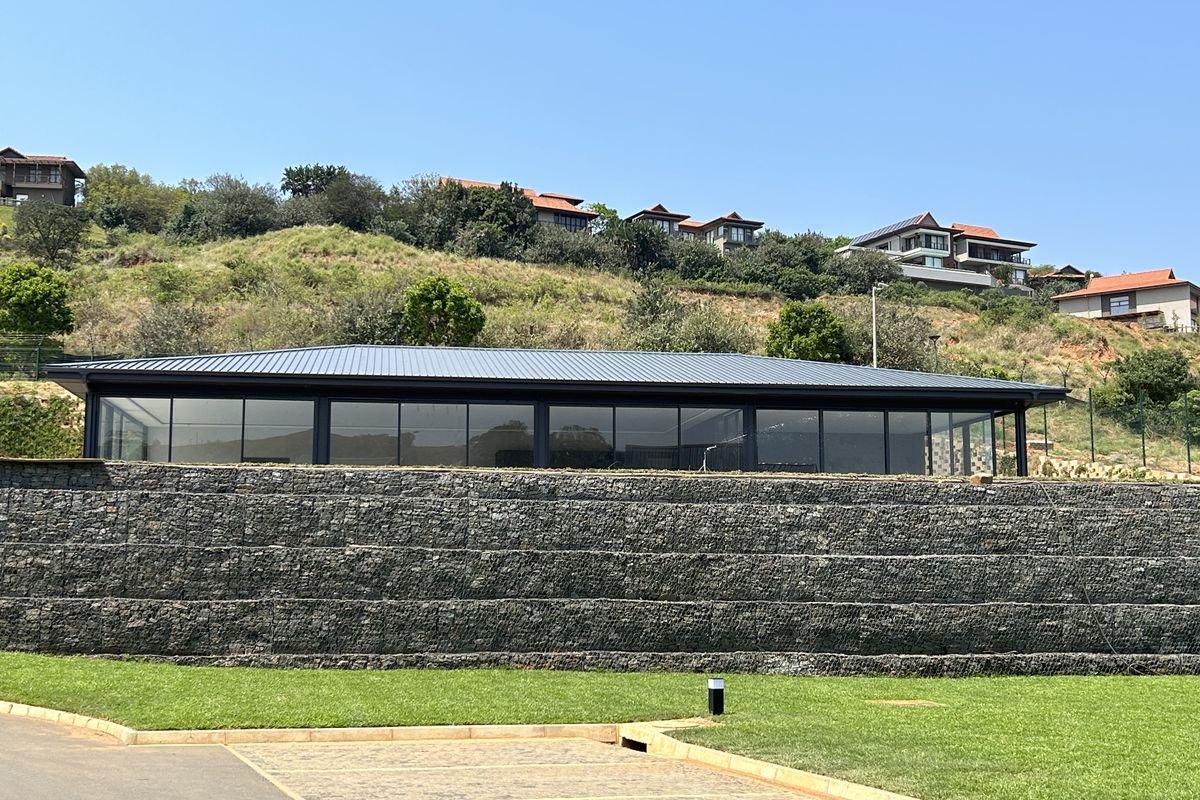 A modern building exterior featuring a large glass facade, a metal roof, and a substantial stone gabion retaining wall set against a hillside.