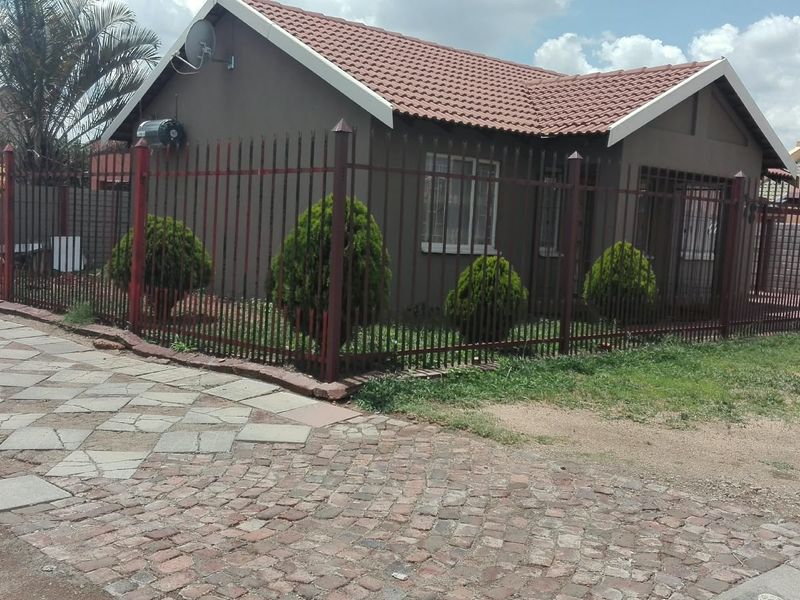 Front exterior view of a single-story house featuring a tiled roof, metal perimeter fencing, and a paved driveway area.