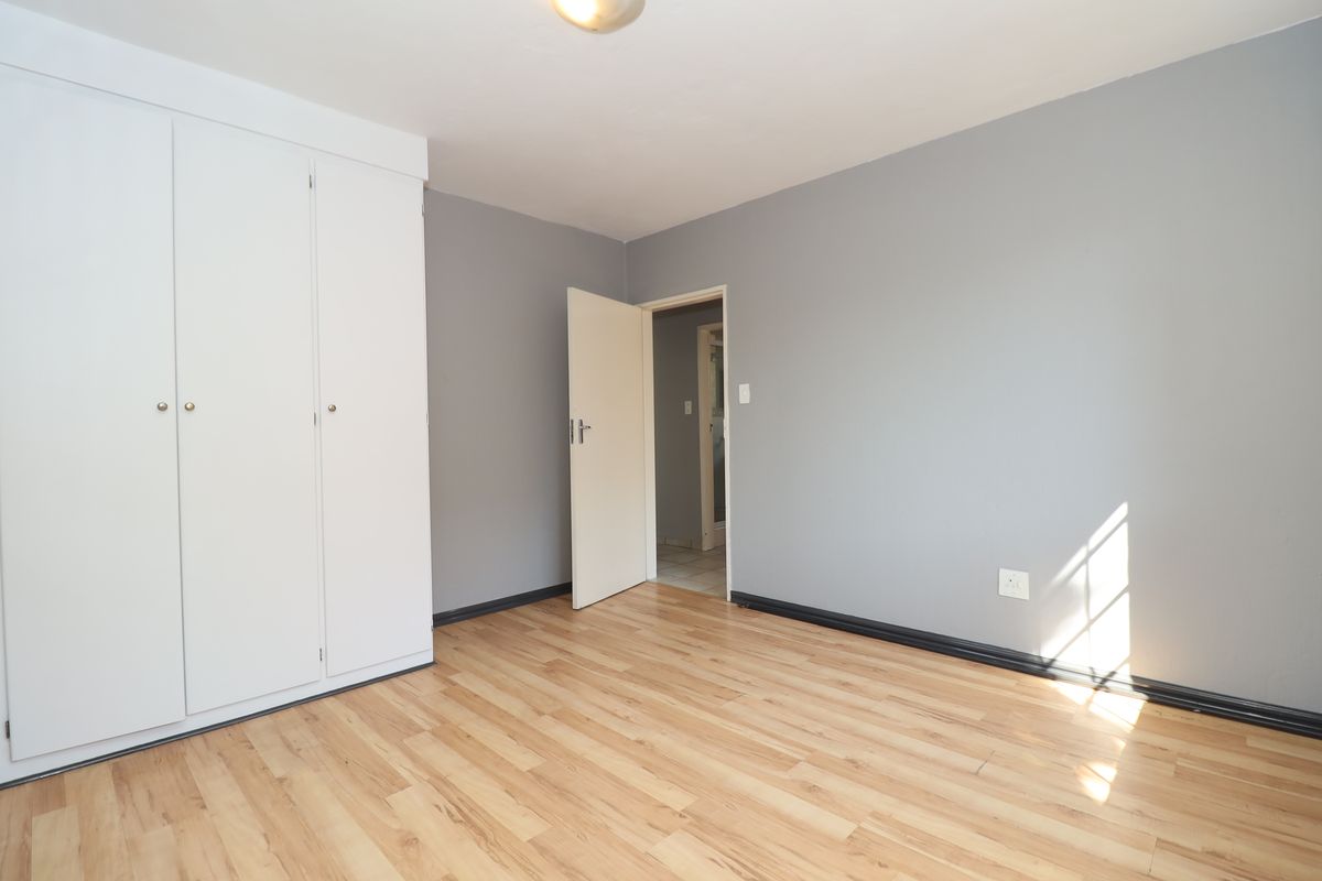 A bright, empty bedroom featuring light wood-look laminate flooring, grey walls, and a built-in white wardrobe.