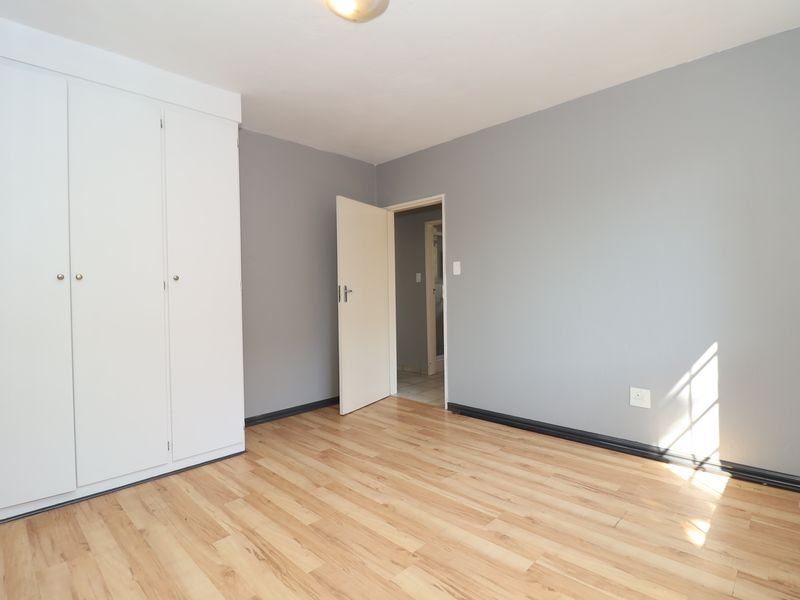 A bright, empty bedroom featuring light wood-look laminate flooring, grey walls, and a built-in white wardrobe.