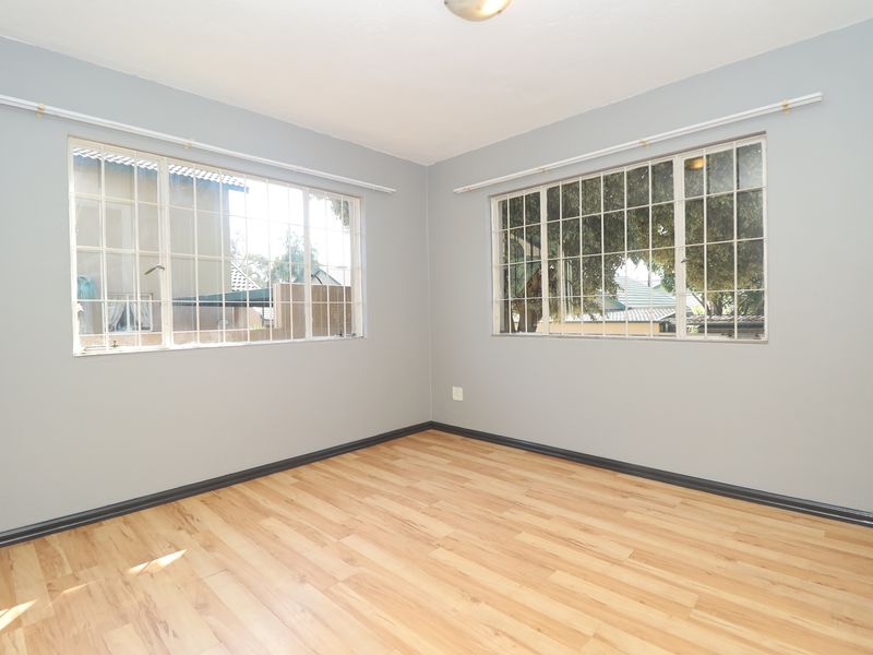 A bright and empty bedroom featuring light wood-look laminate flooring, neutral grey walls, and large windows fitted with security burglar bars.