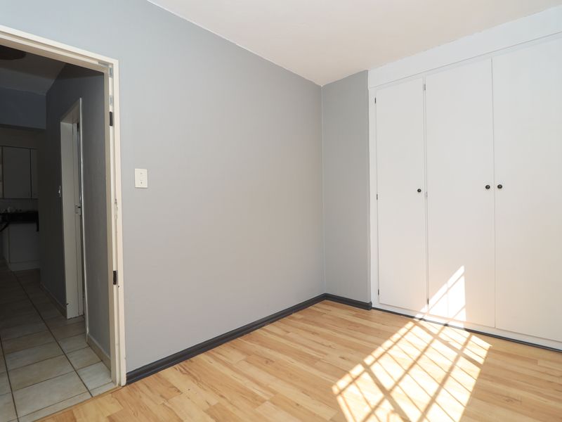 A bright, empty bedroom featuring light wood-look laminate flooring, grey walls, and white built-in cupboards, with a view into the adjacent tiled hallway.