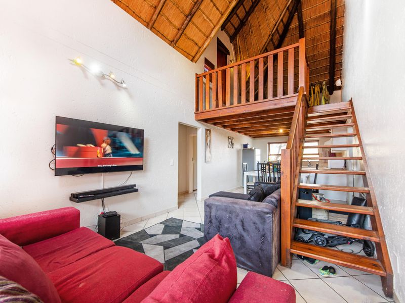 Open-plan living area featuring a double-volume thatch ceiling, exposed timber beams, and a wooden staircase leading to a mezzanine level.