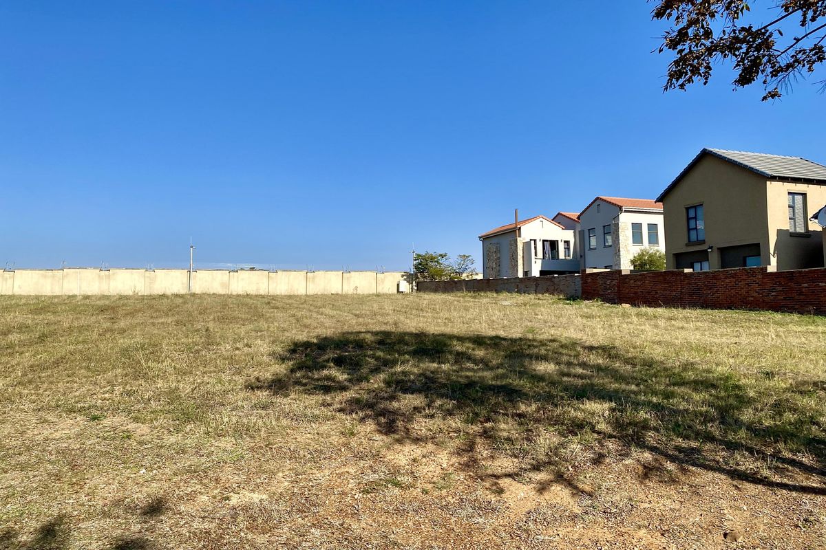A vacant residential plot of land featuring dry grass, enclosed by a perimeter wall, situated in a developed neighborhood.