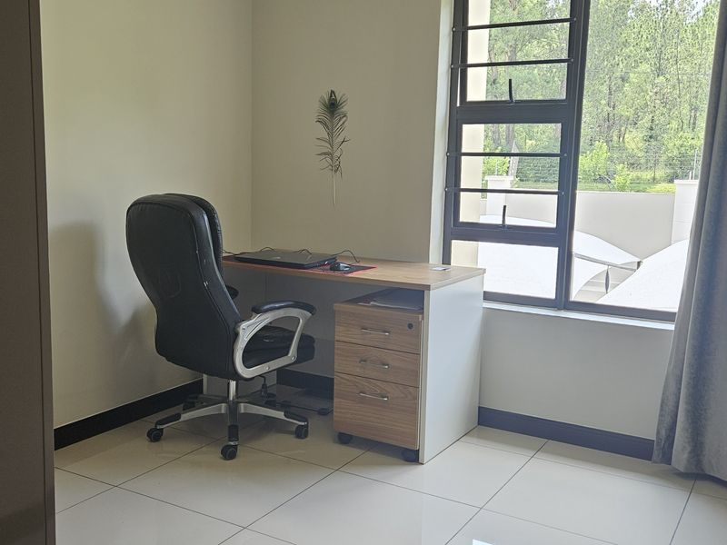 A functional home office space featuring tiled flooring and a large window with security bars, providing natural light and a view of the exterior.