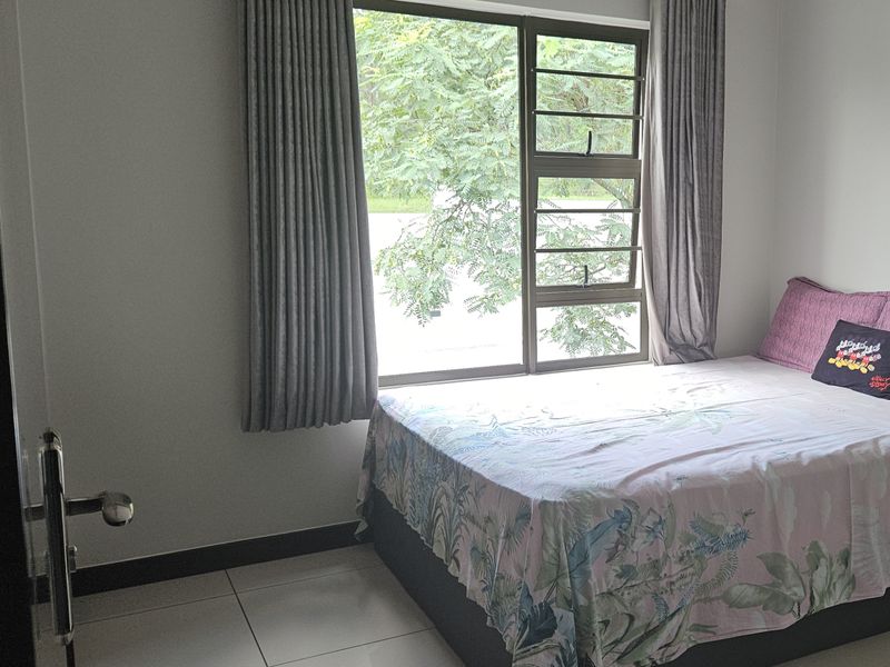 A well-maintained bedroom featuring neutral tiled flooring, clean wall finishes, and a large window with integrated security bars.