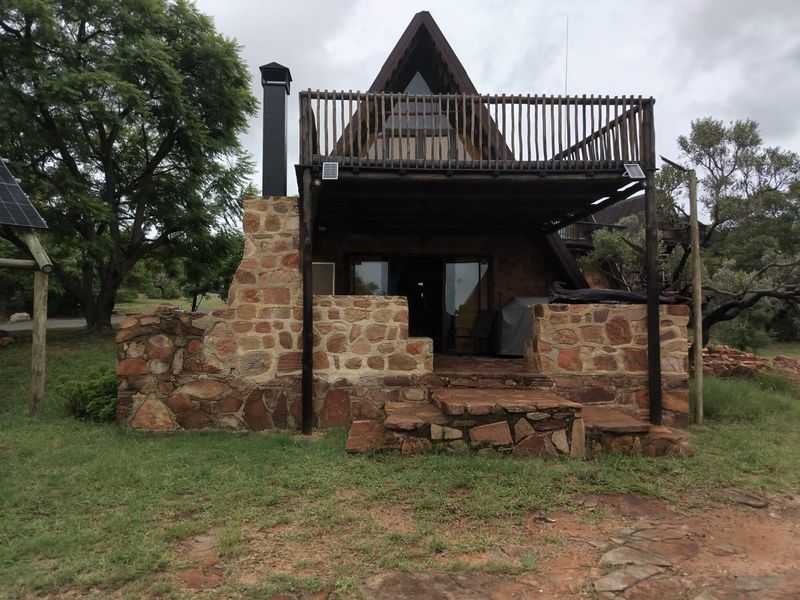 A rustic A-frame house featuring stone masonry, an external fireplace chimney, and an elevated timber balcony overlooking the surrounding garden area.