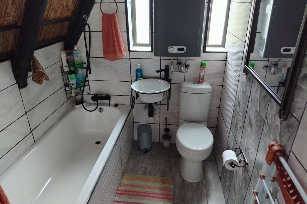 Compact bathroom featuring a bathtub, wall-mounted basin, and toilet, finished with tiled surfaces and a unique exposed thatch roof ceiling.