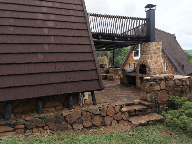 Outdoor patio area featuring stone masonry, a built-in braai with chimney, stone paving, and a timber balcony overhead.