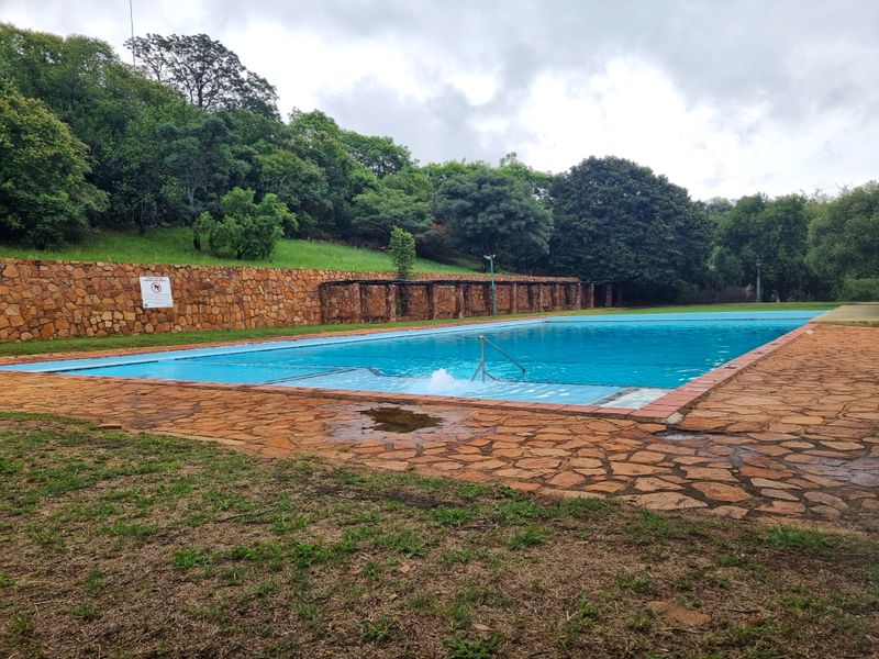 Large outdoor swimming pool area featuring a stone retaining wall, paved surround, and a metal pergola structure set against a landscaped garden backdrop.